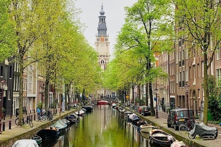 Canal view with boats, lined with trees and buildings, clock tower in distance on a cloudy day.