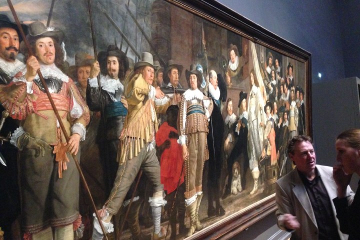 People viewing a large historical painting in a museum, depicting figures in traditional attire.