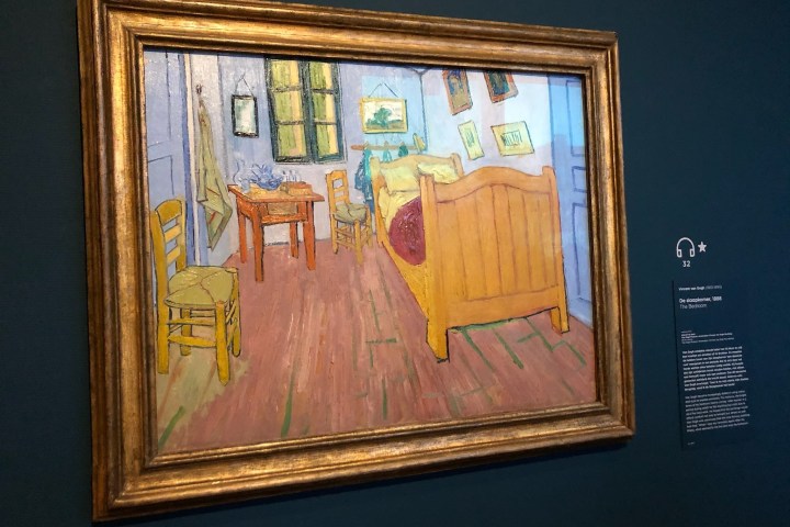Framed painting of a bedroom with wooden furniture and blue walls displayed in a museum.