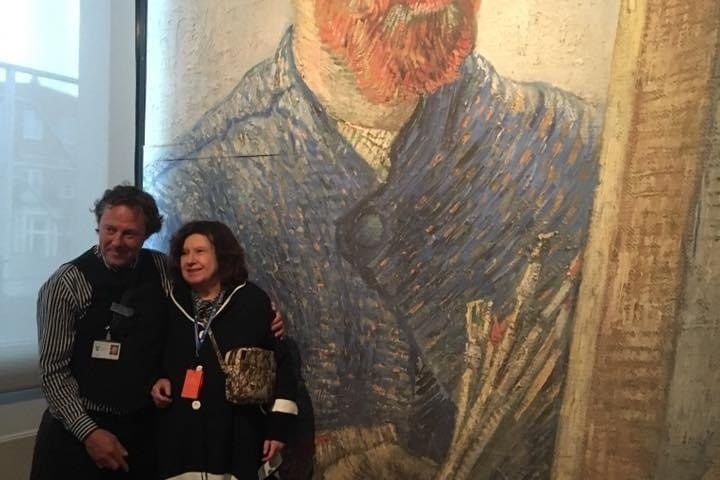 Two people posing in front of a large painting of an artist holding brushes.