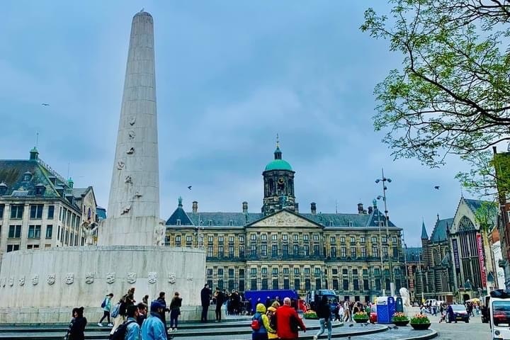 City square with monument, historic building, and people walking on a cloudy day.
