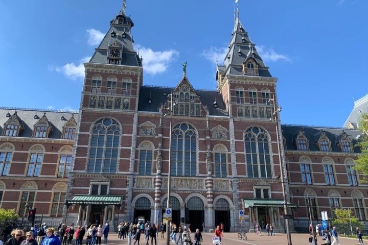 Large brick building with arched windows and towers, people walking in front under a clear blue sky.