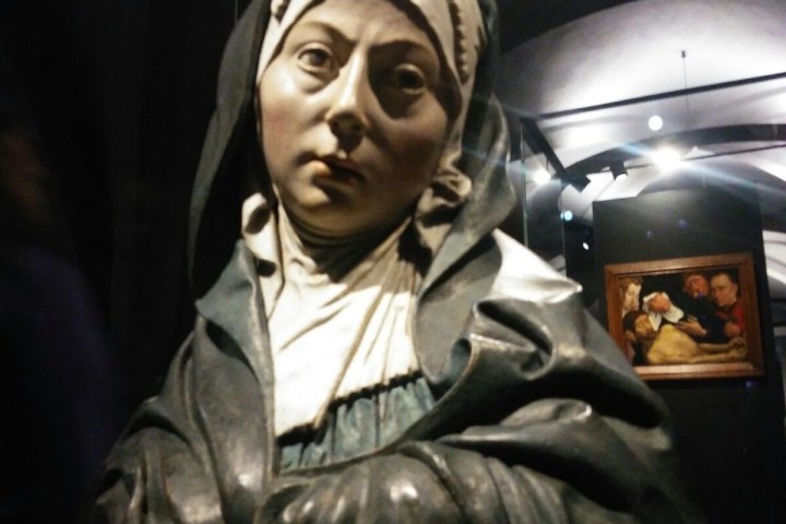 Sculpture bust of a person wearing a hood, with a painting in the background.