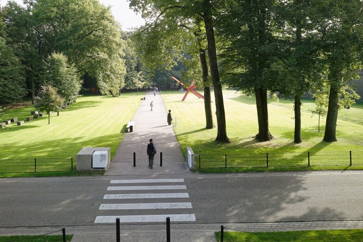 Park with a pathway, trees, and a red sculpture, viewed from a zebra crossing.