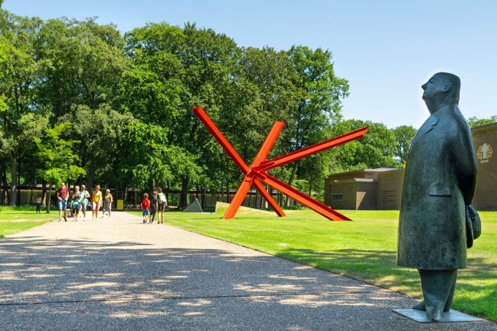 People walking on a path near a statue and red abstract sculpture in a park.