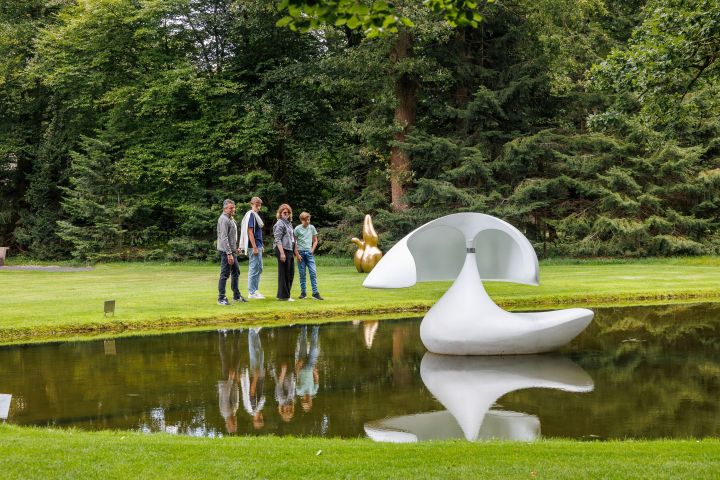 Four people stand by a pond with abstract sculptures amid trees.
