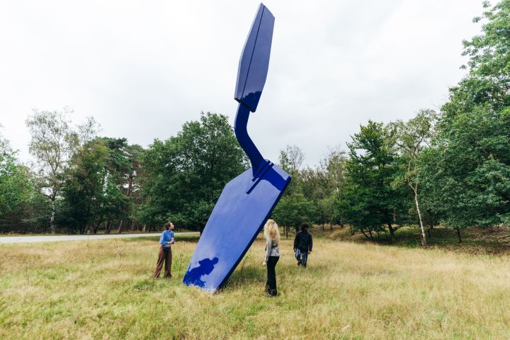 Three people stand near a large, abstract blue sculpture in a grassy field with trees.