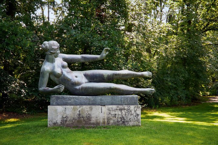 Reclining nude statue on a pedestal in a lush park setting.